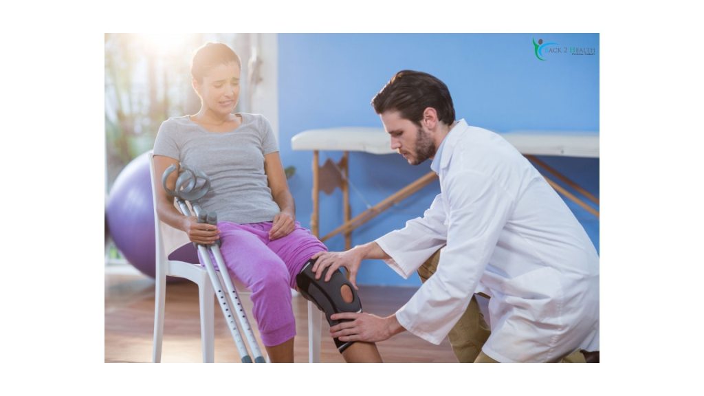 A male physical therapist in a white coat fitting a supportive knee brace on a female patient holding crutches at Back 2 Health Physical Therapy.