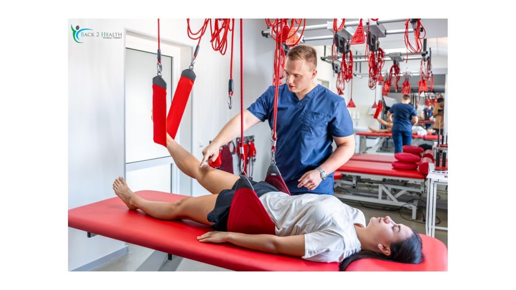 A physical therapist at Back 2 Health guides a patient through a rehabilitation session using a Redcord suspension system. The patient lies on a red treatment table while her legs are supported by red slings, allowing for controlled, low-impact movement to improve core stability and joint function.