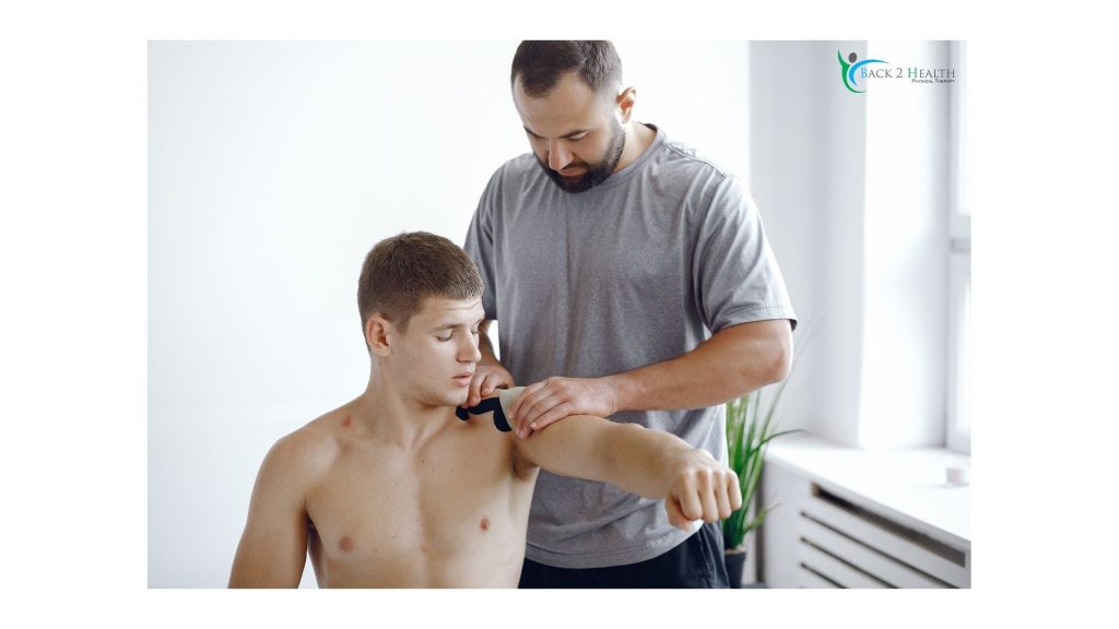 A physical therapist at Back 2 Health applies kinesiology tape to the shoulder of a young male athlete. The therapist is carefully positioning the black tape to provide joint stability and support during movement, while the patient holds his arm extended to assist in the application process.