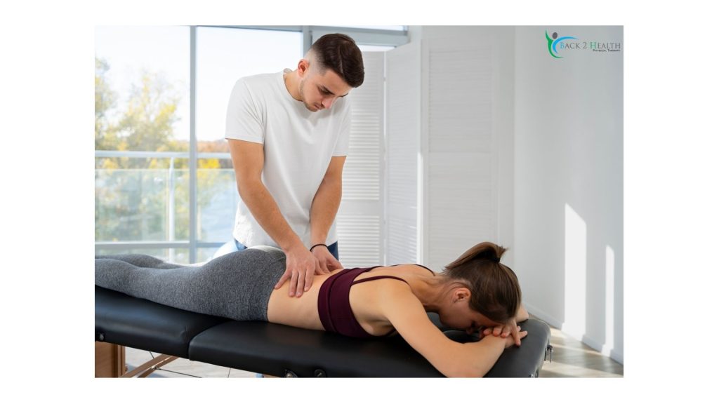 A male physical therapist at Back 2 Health performs manual therapy on a female patient lying face down on a clinical treatment table. The therapist is wearing a white T-shirt and gently applying pressure to the patient’s lower back to treat stiffness and pain. The patient is wearing grey leggings and a dark purple sports bra. The bright, modern clinic features large windows in the background showing outdoor trees.