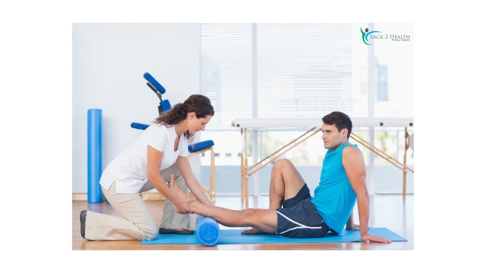 A female physical therapist kneeling on a blue exercise mat, assisting a male patient as he rolls his calf muscle over a blue foam roller in a bright clinic setting.